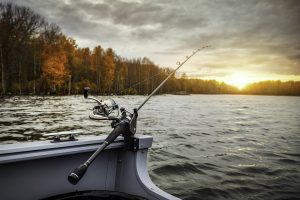 A fishing rod connected to the edge of a boat on a lake with trees and the sun setting in the background. | Fishing around Fort Smith, AR.