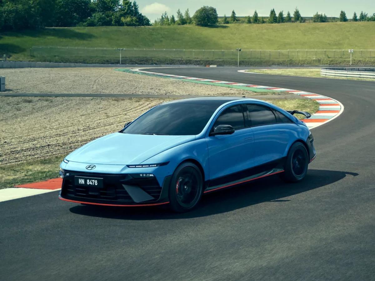 Front-quarter view of a Performance Blue 2026 Hyundai IONIQ 6 N cornering on a racetrack with its aggressive front fascia and wide-body stance visible.