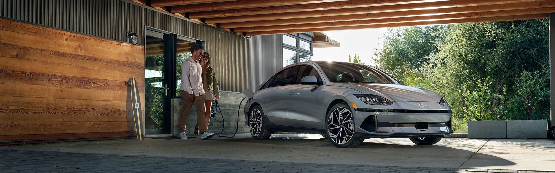 A couple stands at the door of their modern home while their silver car charges under a wooden beam carport.